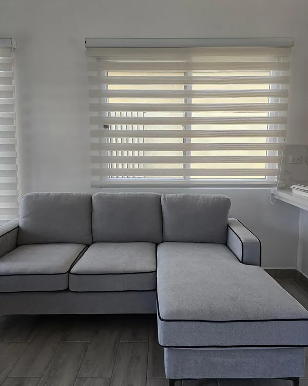 Cortina Dual Shade blanca sobre la ventana de una sala, detrás de un sofá gris en L