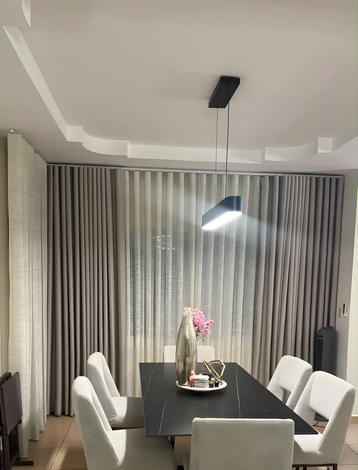 Cortinas Ripple Fold grises a los lados de un velo de lino, de suelo a techo, detrás de un comedor con mesa negra y sillas blancas
